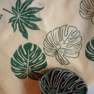 May include: A close-up of a fabric with green leaf prints. The fabric has a light beige background and features several different leaf designs, including a large monstera leaf, a smaller monstera leaf, and a smaller palm leaf. The fabric is likely used for crafting or home decor.