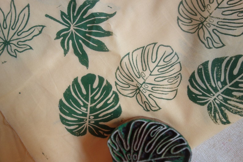 May include: A close-up of a fabric with green leaf prints. The fabric has a light beige background and features several different leaf designs, including a large monstera leaf, a smaller monstera leaf, and a smaller palm leaf. The fabric is likely used for crafting or home decor.