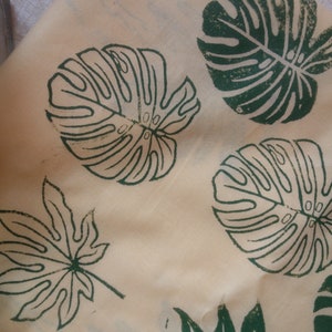 May include: A close-up of a beige fabric with green leaf prints. The leaves are in a variety of shapes and sizes, including a large monstera leaf and a smaller maple leaf.