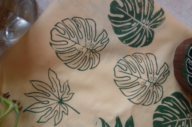 May include: A close-up of a beige fabric with green leaf prints. The leaves are in a variety of shapes and sizes, including a large monstera leaf and a smaller maple leaf.