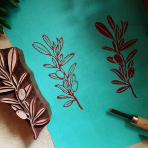 Puede incluir: Un bloque de madera tallada con un dise&ntilde;o de rama de olivo, colocado junto a una hoja de papel color verde azulado que muestra dos impresiones en tinta roja del mismo patr&oacute;n, junto a dos herramientas de tallado de metal con mangos de madera.