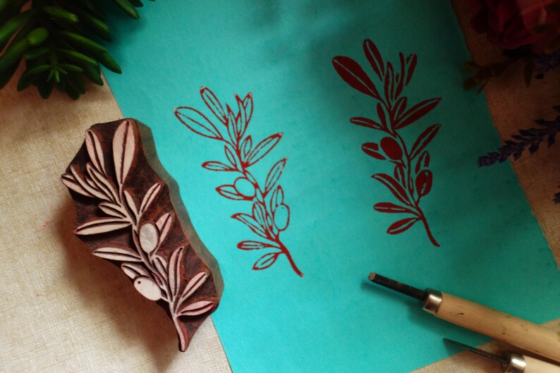 Puede incluir: Un bloque de madera tallada con un dise&ntilde;o de rama de olivo, colocado junto a una hoja de papel color verde azulado que muestra dos impresiones en tinta roja del mismo patr&oacute;n, junto a dos herramientas de tallado de metal con mangos de madera.
