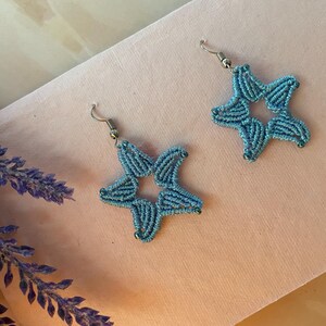 May include: A pair of light blue star-shaped earrings crafted from intricate thread work. Each earring features a five-pointed star design with a hollow center and is attached to a silver-toned metal hook for hanging, showcasing detailed fiber art.