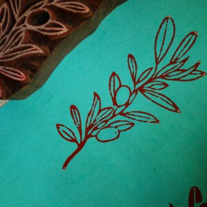 Puede incluir: Un bloque de madera tallado con un dise&ntilde;o de rama de olivo colocado junto a una superficie de papel verde azulado con un patr&oacute;n estampado de rama de olivo rojo oscuro. El bloque muestra la talla en relieve utilizada para el motivo bot&aacute;nico.
