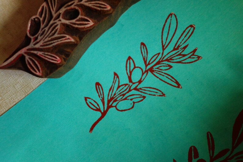 Puede incluir: Un bloque de madera tallado con un dise&ntilde;o de rama de olivo colocado junto a una superficie de papel verde azulado con un patr&oacute;n estampado de rama de olivo rojo oscuro. El bloque muestra la talla en relieve utilizada para el motivo bot&aacute;nico.