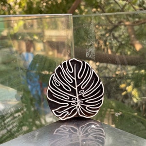 May include: A black wooden block carving of a white monstera leaf. The carving is on a glass surface.