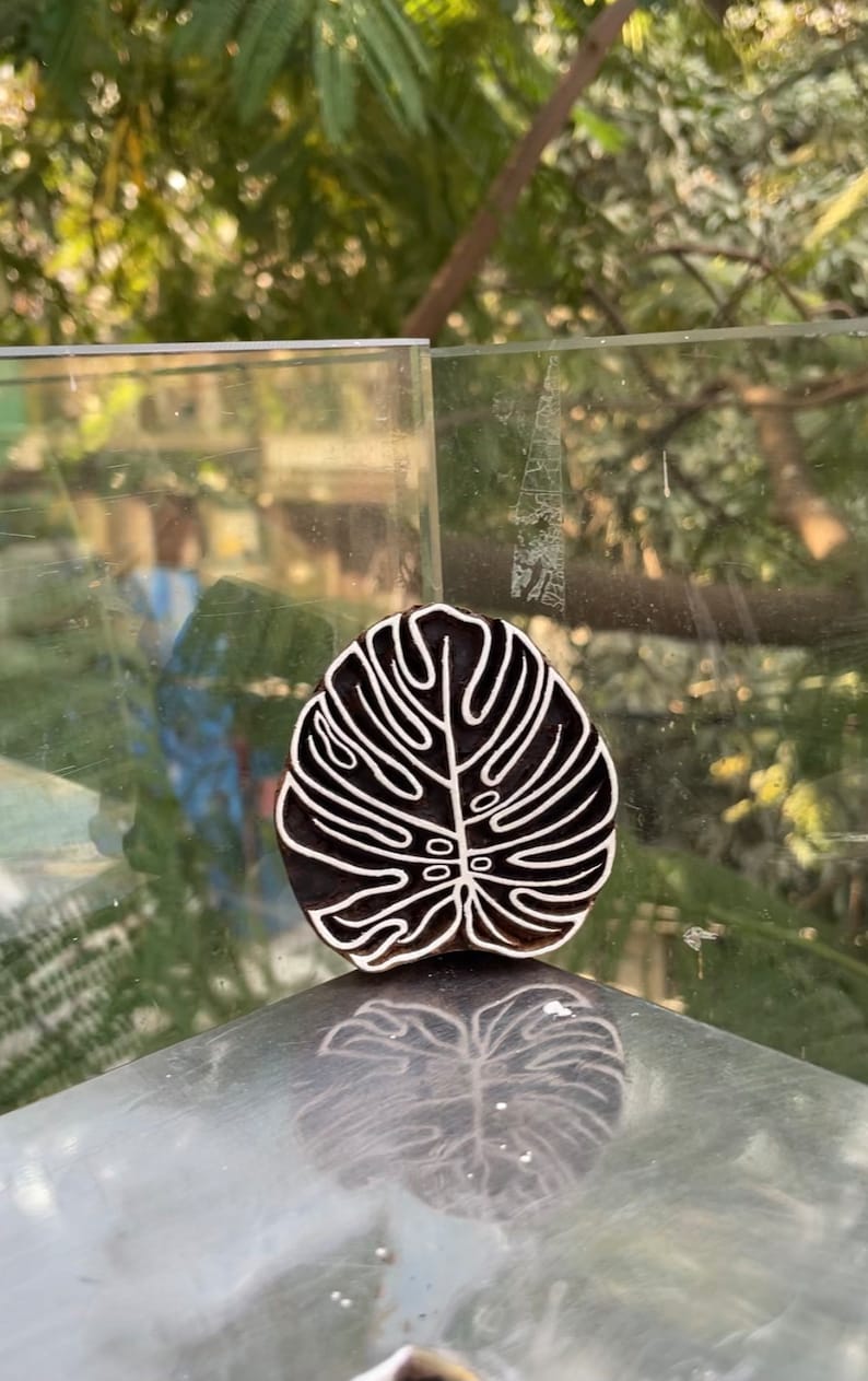 May include: A black wooden block carving of a white monstera leaf. The carving is on a glass surface.
