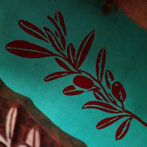 Puede incluir: Una superficie de tela de color turquesa vibrante presenta un dise&ntilde;o impreso en rojo oscuro de una rama de olivo con hojas y dos aceitunas. Junto a la tela, se ve una parte de un bloque de madera tallada con un patr&oacute;n de rama de olivo.