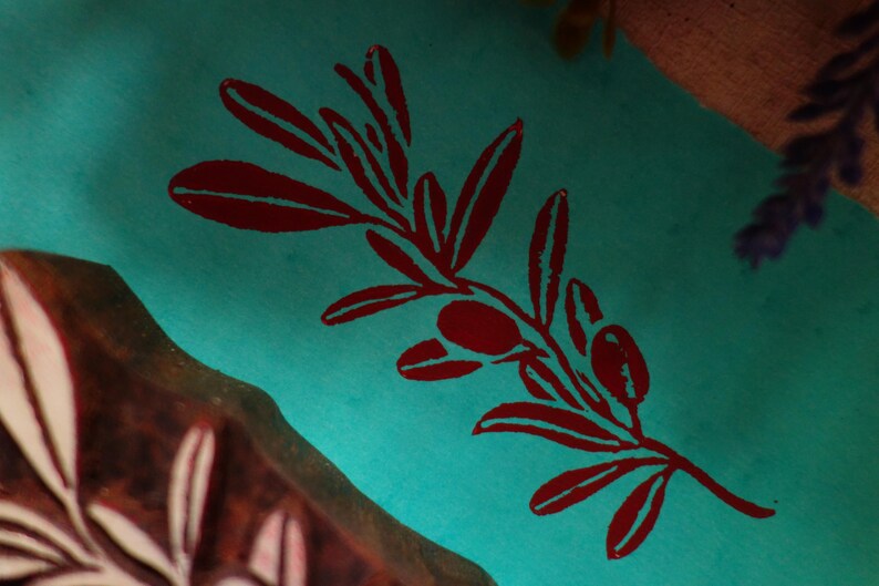 Puede incluir: Una superficie de tela de color turquesa vibrante presenta un dise&ntilde;o impreso en rojo oscuro de una rama de olivo con hojas y dos aceitunas. Junto a la tela, se ve una parte de un bloque de madera tallada con un patr&oacute;n de rama de olivo.
