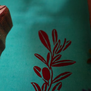 Puede incluir: Un sello de madera marr&oacute;n oscuro con un patr&oacute;n de hojas tallado junto a una superficie de papel color verde azulado con un dise&ntilde;o estampado en tinta roja de una rama frondosa con un fruto ovalado, mostrando la impresi&oacute;n clara del sello.