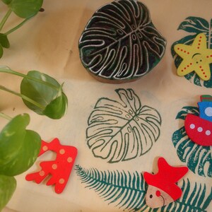 May include: A collection of wooden stamps and toys with a tropical theme. The stamps are carved with a monstera leaf design and the toys are painted in bright colors, including red, yellow, blue, and green. The toys include a starfish, a boat, and a fish.