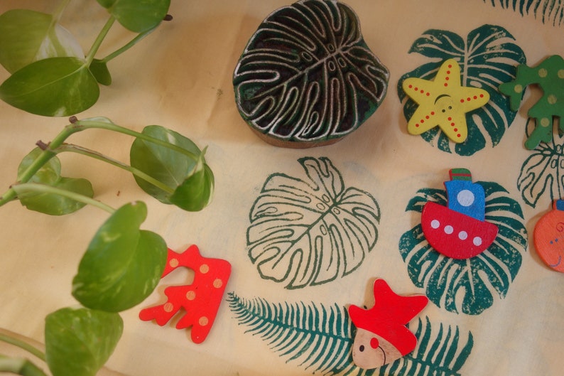 May include: A collection of wooden stamps and toys with a tropical theme. The stamps are carved with a monstera leaf design and the toys are painted in bright colors, including red, yellow, blue, and green. The toys include a starfish, a boat, and a fish.