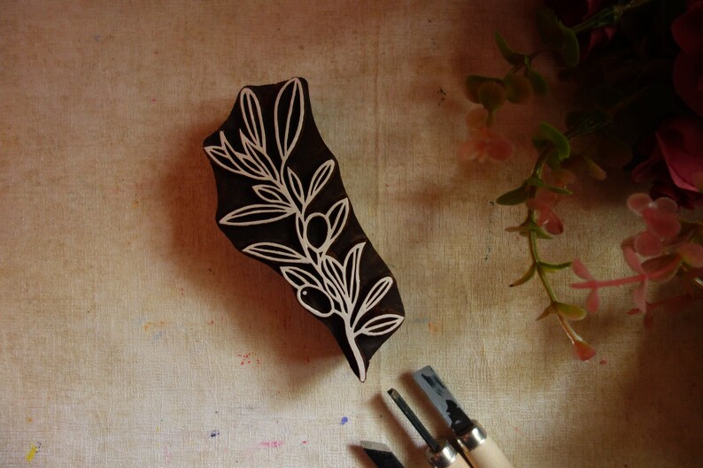 Puede incluir: Un sello de madera de color marr&oacute;n oscuro para impresi&oacute;n con un dise&ntilde;o tallado de rama de olivo con hojas y dos aceitunas. El sello tiene una forma org&aacute;nica irregular y est&aacute; colocado junto a dos peque&ntilde;as herramientas de tallado de metal.