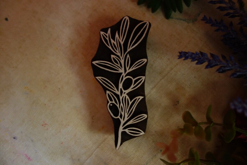 Puede incluir: Un bloque de impresi&oacute;n de madera marr&oacute;n oscuro con un dise&ntilde;o tallado de una rama de olivo con hojas y dos aceitunas. Las l&iacute;neas talladas son de color claro, creando un patr&oacute;n bot&aacute;nico de alto contraste en la superficie del bloque de madera.