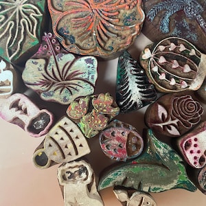 May include: A collection of assorted wooden printing blocks featuring various carved designs including flowers, leaves, a rose, a tree, and geometric shapes. The blocks show remnants of colorful paint, including green, red, and blue, on their textured surfaces.