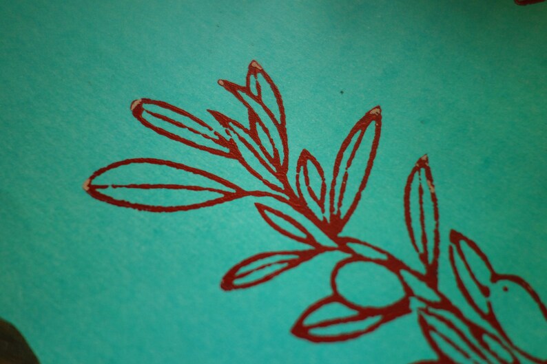 Puede incluir: Una superficie de papel turquesa vibrante con una impresi&oacute;n de tinta roja oscura de una rama bot&aacute;nica. La ilustraci&oacute;n muestra un tallo central con m&uacute;ltiples hojas alargadas y un peque&ntilde;o fruto redondo, representado en un estilo de l&iacute;nea audaz y texturizado.