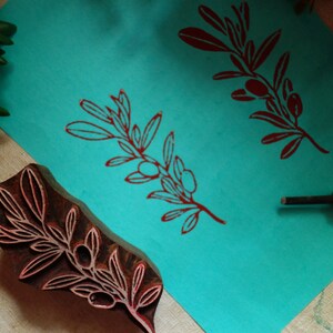 Puede incluir: Un sello de bloque de madera marr&oacute;n oscuro con un dise&ntilde;o de rama de olivo, colocado junto a una hoja de papel turquesa brillante que muestra dos impresiones de tinta roja del mismo motivo, acompa&ntilde;ado de una herramienta de tallado con punta met&aacute;lica.