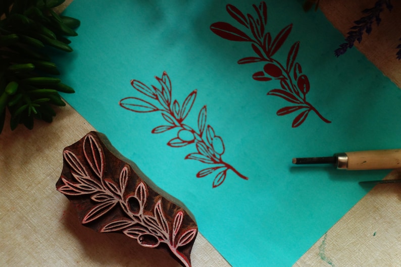 Puede incluir: Un sello de bloque de madera marr&oacute;n oscuro con un dise&ntilde;o de rama de olivo, colocado junto a una hoja de papel turquesa brillante que muestra dos impresiones de tinta roja del mismo motivo, acompa&ntilde;ado de una herramienta de tallado con punta met&aacute;lica.