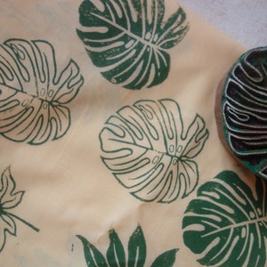 May include: A close-up of a fabric with green leaf prints. The fabric has a light beige background and features several different leaf designs, including a monstera leaf, a maple leaf, and a fern leaf. The leaves are printed in a dark green color.