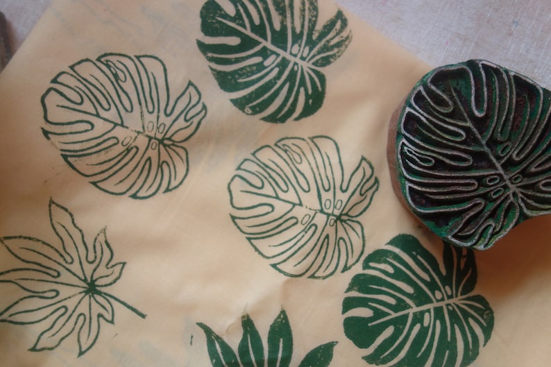 May include: A close-up of a fabric with green leaf prints. The fabric has a light beige background and features several different leaf designs, including a monstera leaf, a maple leaf, and a fern leaf. The leaves are printed in a dark green color.