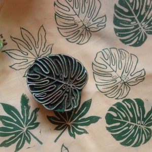 May include: A close-up of a fabric with green leaf prints. The fabric is a light beige color. There is a dark wooden block with a leaf design on it.