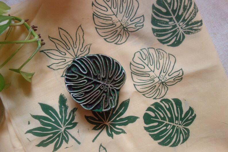 May include: A close-up of a fabric with green leaf prints. The fabric is a light beige color. There is a dark wooden block with a leaf design on it.