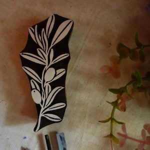 Puede incluir: Un bloque de impresi&oacute;n de madera marr&oacute;n oscuro con un dise&ntilde;o tallado de una rama de olivo con hojas y dos aceitunas. Las &aacute;reas talladas son de color claro, creando un patr&oacute;n de alto contraste sobre la superficie de madera oscura del bloque.