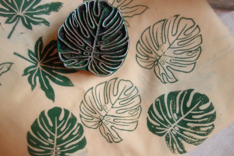 May include: A close-up of a green leaf-shaped rubber stamp on a beige surface. The stamp is surrounded by several green leaf prints.