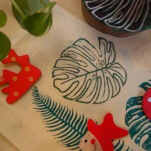 May include: A collection of wooden stamps featuring a variety of designs, including a monstera leaf, a fern, a starfish, a boat, and a coral. The stamps are painted in bright colors, including red, yellow, blue, and green.