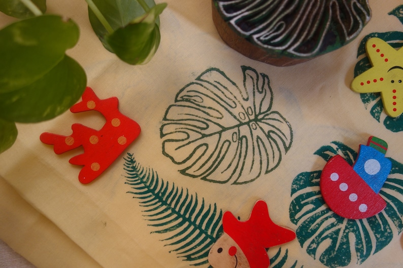 May include: A collection of wooden stamps featuring a variety of designs, including a monstera leaf, a fern, a starfish, a boat, and a coral. The stamps are painted in bright colors, including red, yellow, blue, and green.