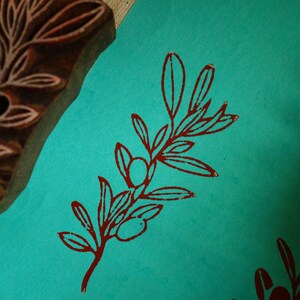 Puede incluir: Un bloque de madera tallada con un dise&ntilde;o de rama de olivo junto a una superficie de papel color verde azulado con una impresi&oacute;n estampada de rama de olivo en rojo oscuro. El sello muestra el patr&oacute;n en relieve usado para la ilustraci&oacute;n.