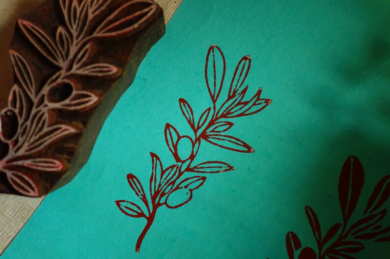 Puede incluir: Un bloque de madera tallada con un dise&ntilde;o de rama de olivo junto a una superficie de papel color verde azulado con una impresi&oacute;n estampada de rama de olivo en rojo oscuro. El sello muestra el patr&oacute;n en relieve usado para la ilustraci&oacute;n.
