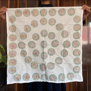 以下が含まれることがあります： ピンクと黄色の花の模様が緑の葉で囲まれた白い生地。 花柄は生地全体に均等に配置されています。 生地は人が持っています。