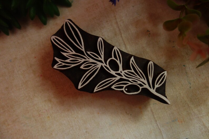 Puede incluir: Un sello de madera de color marr&oacute;n oscuro con un dise&ntilde;o tallado de una rama de olivo con hojas y dos aceitunas. Las l&iacute;neas talladas son de color claro, creando un alto contraste con la superficie de madera oscura del bloque rectangular.