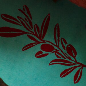 Puede incluir: Una superficie de color verde azulado vibrante presenta un gr&aacute;fico serigrafiado de color rojo oscuro de una rama de olivo. El dise&ntilde;o muestra hojas detalladas y dos peque&ntilde;as aceitunas dispuestas a lo largo de un tallo central con contraste.