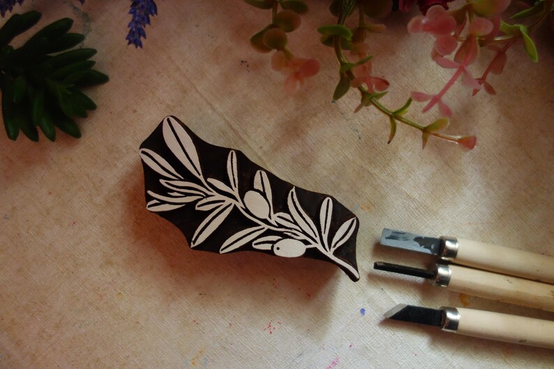 Puede incluir: Un sello de madera de color marr&oacute;n oscuro para impresi&oacute;n con un dise&ntilde;o tallado en blanco de una rama de olivo con hojas y dos aceitunas. Tres herramientas de tallado de metal con mangos de madera clara se sit&uacute;an a la derecha del bloque.
