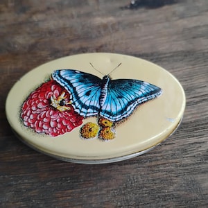 May include: An oval, yellow tin with a butterfly and flower design. The butterfly has blue and black wings, and the flower is red. The tin is sitting on a wooden surface.