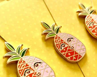 May include: Close-up of several pineapple-shaped enamel pins with gold trim. The pins are divided into red and pink halves, with a face design. The green leaves and gold stems add detail. The pins are on yellow paper.
