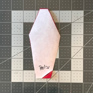 May include: A red and white paper craft with the word "Peel" written on it. The paper is folded over a red surface, revealing the red color underneath. The craft is on a gray and black checkered surface.