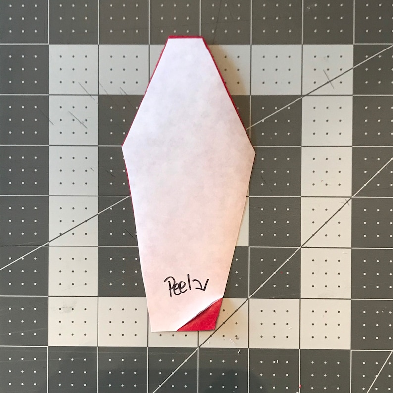 May include: A red and white paper craft with the word "Peel" written on it. The paper is folded over a red surface, revealing the red color underneath. The craft is on a gray and black checkered surface.
