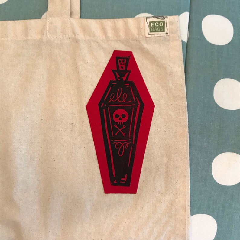May include: A cream-colored tote bag with a red coffin-shaped graphic. The graphic features a black skull and crossbones design. The bag has a small tag that reads "ECO BAGS".