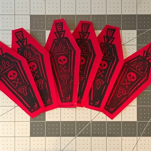 May include: A collection of six red coffin-shaped paper cutouts. Each cutout features a black illustration of a coffin with a skull and crossbones, a decorative bottle top, and the word "ele".