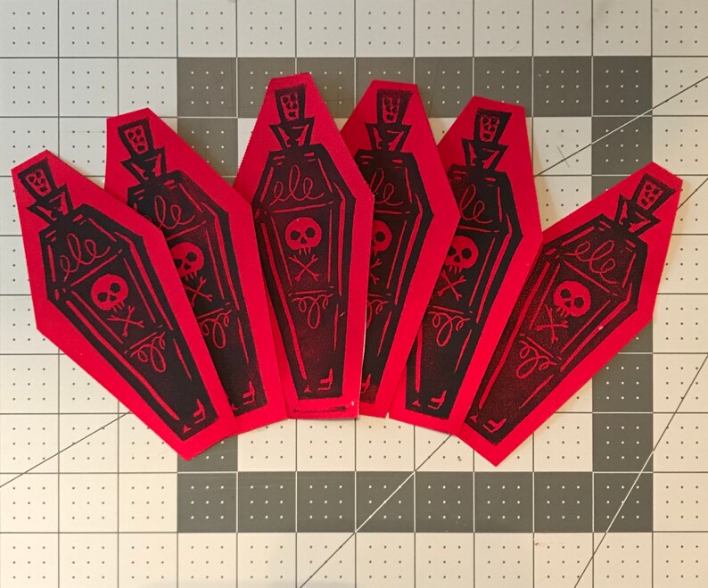 May include: A collection of six red coffin-shaped paper cutouts. Each cutout features a black illustration of a coffin with a skull and crossbones, a decorative bottle top, and the word "ele".