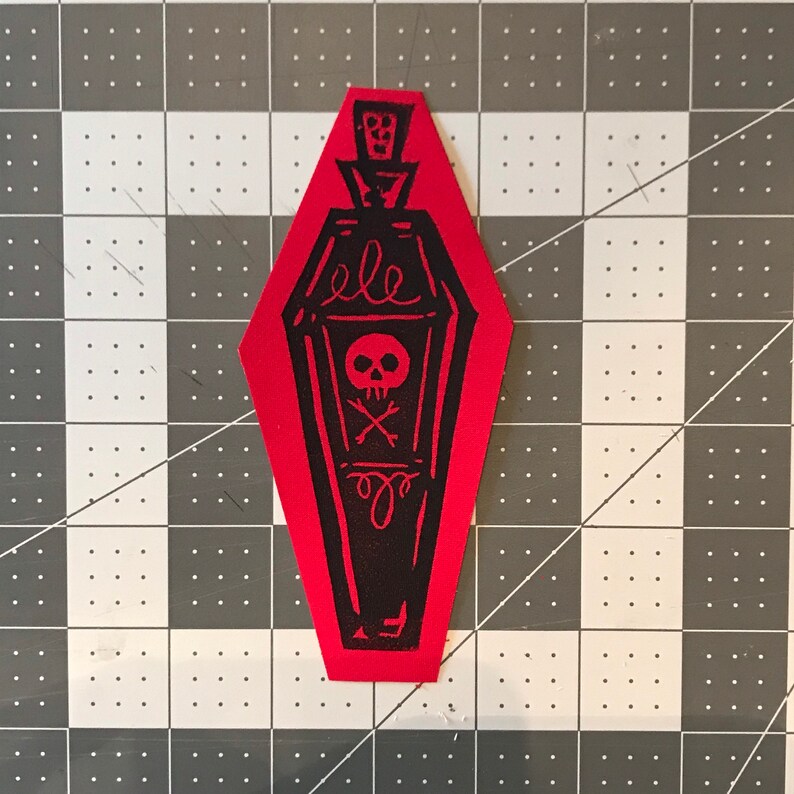 May include: A red, coffin-shaped patch with a black illustration of a coffin-shaped bottle. The bottle design includes a skull and crossbones, and the word "ele". The patch is on a black and white checkered surface.