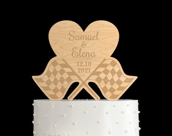 Könnte beinhalten: Holz-Tortenaufsatz für Hochzeit mit Herzform und zwei karierten Flaggen. Das Herz ist mit den Namen "Samuel & Elena" und dem Datum "12.10.2021" graviert.