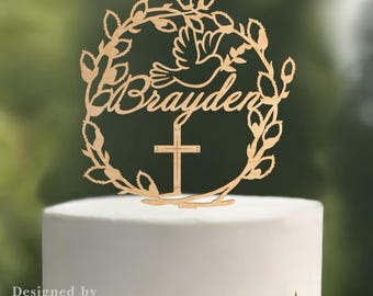 May include: A wooden cake topper featuring the name "Brayden" in script, encircled by a wreath of leaves and a dove. A cross is centered below the name. The topper is placed on a white cake with a white rose.