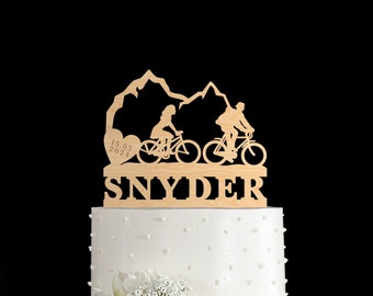 May include: A wooden wedding cake topper featuring a couple riding bicycles with mountains in the background. The topper also includes a heart with the date "15.03.2023" and the last name "SNYDER".
