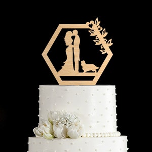 May include: Wooden wedding cake topper featuring a couple kissing, a dog, and a leafy design with hearts. The topper is in a hexagon shape and is made of natural wood.