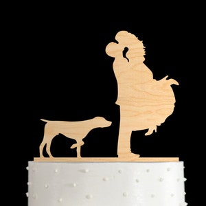 May include: A white wedding cake with a wooden topper of a couple kissing and a dog standing in front of them. The cake is decorated with white frosting and white polka dots.