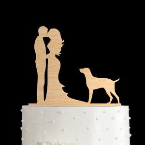 May include: A white wedding cake with a wooden topper of a couple kissing and a dog standing behind them. The cake is decorated with white frosting and white polka dots.